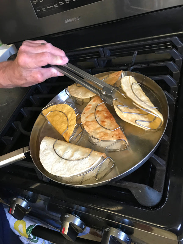 Person using tongs to handle tacos in a frying pan on a stove.