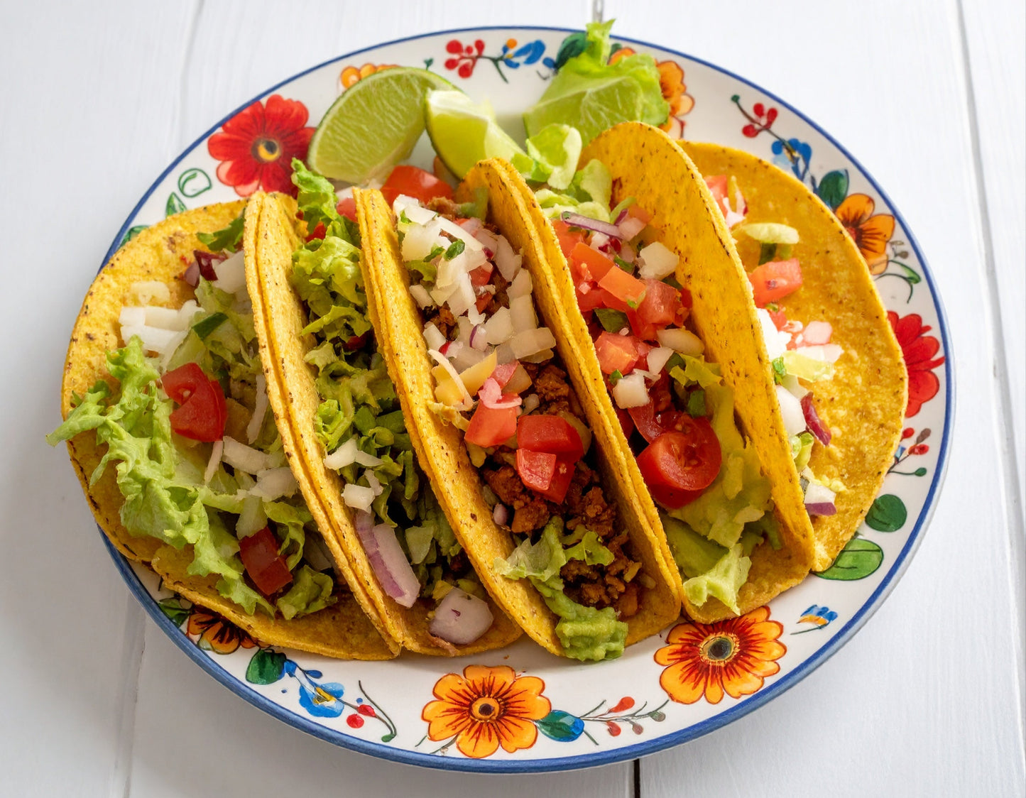Four perfect tacos on a decorative plate made with the Original Mi taco stand, a stainless steel taco shaper. Colorful plate of crispy homemade tacos loaded with lettuce, onions, tomatoes, and lime