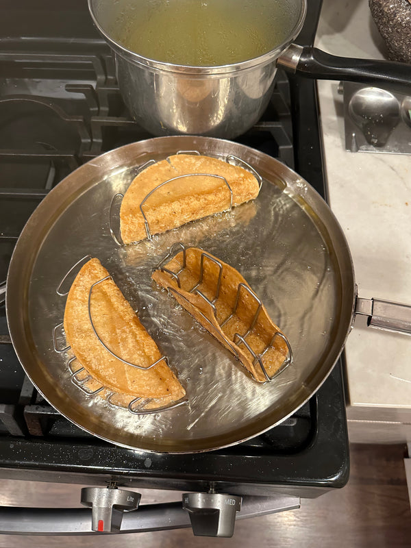 Tortillas frying in oil on stainless steel taco frames in a pan to create crispy taco shells