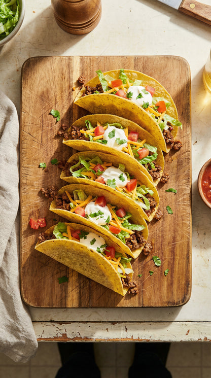 Row of crispy tacos topped with lettuce, tomatoes, cheese, and sour cream on a wooden cutting board
